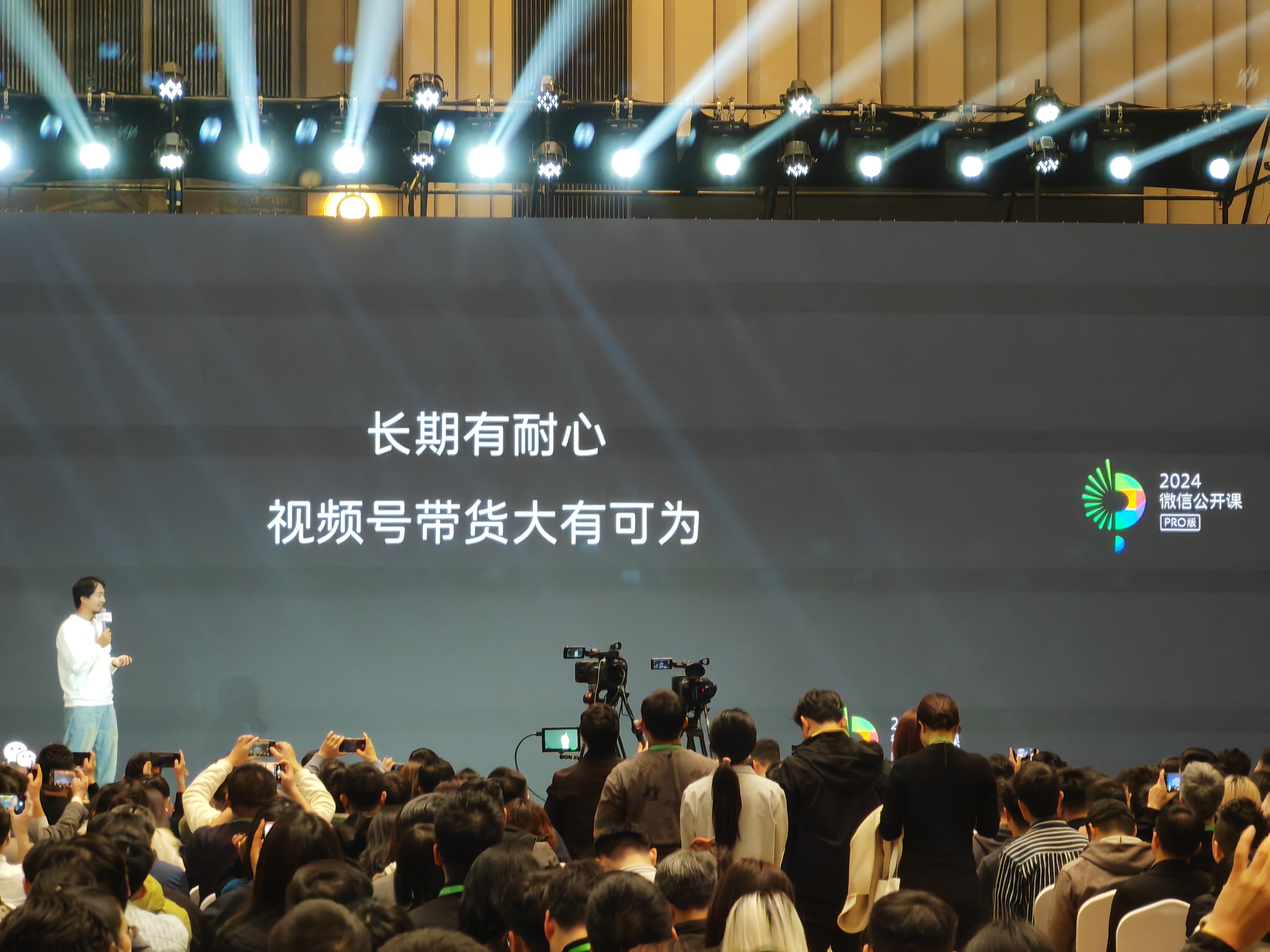 图片展示了2024微信公开课PRO版的现场场景。一位演讲者站在舞台左侧，手持话筒正在讲话。舞台背景大屏上写着“长期有耐心 视频号带货大有可为”，右上角有2024微信公开课PRO版标识。台下聚集着众多观众，部分人举起手机拍照或录像，还有摄像机在进行录制。这张图片与文档中关于2024微信公开课视频号直播带货分享会的内容相呼应，直观呈现了公开课现场氛围。