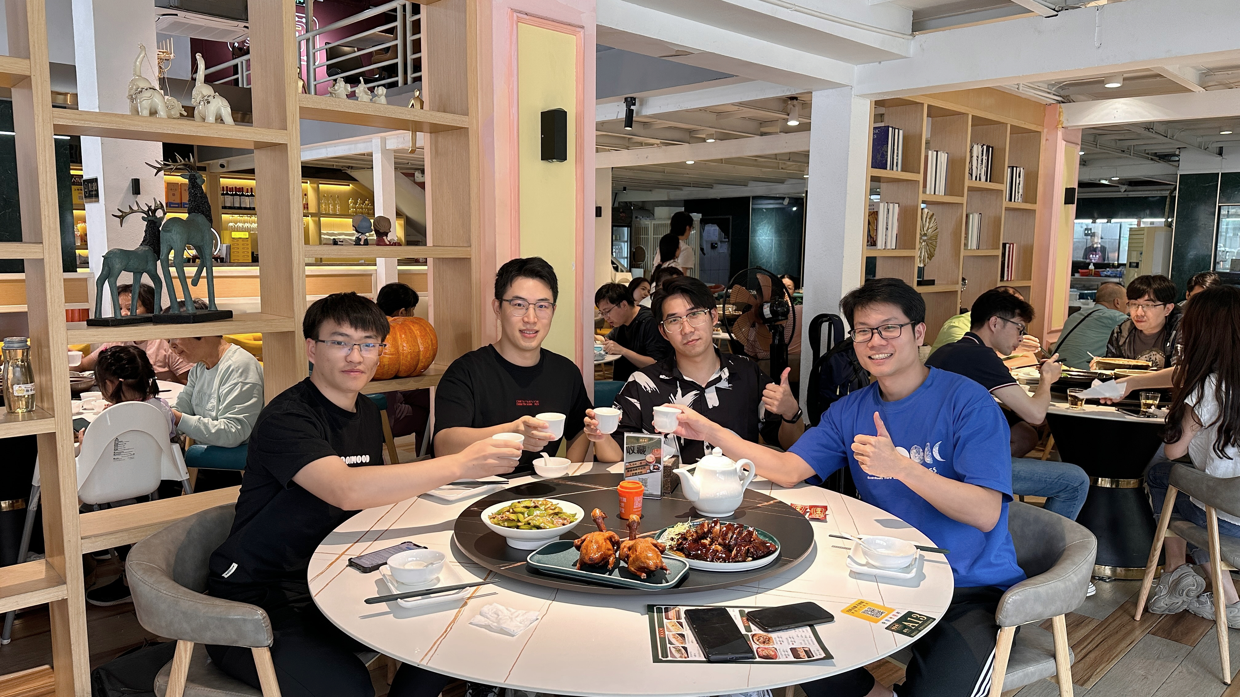 图片展示了一群人在餐厅聚会的场景。四人围坐在圆桌旁，桌上摆放着食物和茶具，其中一人举杯示意，其他人也做出手势。背景中还有其他人在用餐，环境宽敞明亮，有书架、装饰品等。这张图片与文档中“感谢@陈天的引荐，成功签约一家上市公司，让我们有机会向头部企业展示我们的实力”相关，可能是签约后团队庆祝的场景，体现了团队的凝聚力和喜悦之情。