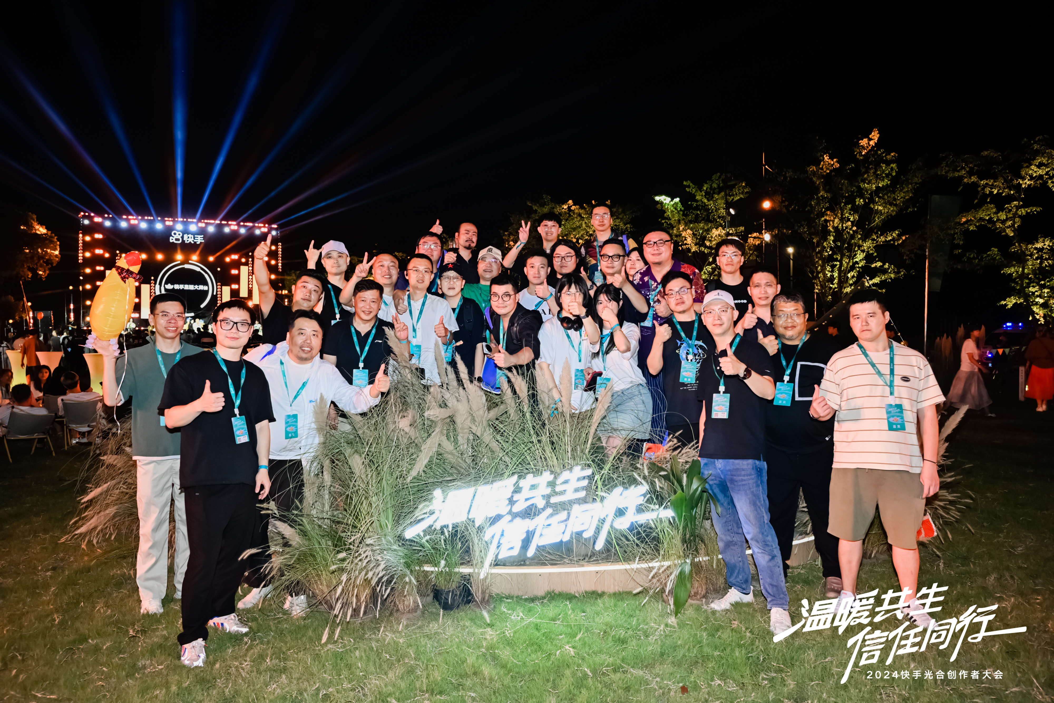 图片展示的是快手光合创作者大会的场景。夜晚，灯光璀璨，背景有“快手光合创作者大会”字样。多人聚集在“温暖共生 信任同行”标识前，面带微笑，有的竖起大拇指，有的比出胜利手势，显得十分开心。部分人佩戴着蓝色证件，背景中还有其他参会者。此图与上文提到的受可灵官方邀请参加快手光合创作者大会相呼应，展现了大会的热闹氛围。