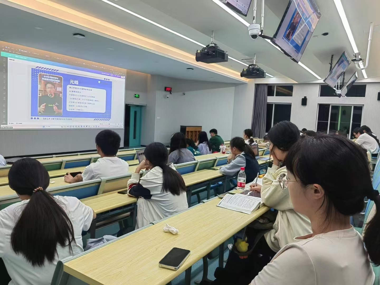 图片展示了一间教室场景，教室前方有学生在认真听讲，前方投影屏幕上显示着“元峰”相关内容，旁边有“AI时代，机会属于勇于尝试的人”等文字。教室里摆放着长桌，桌上有笔记本、水瓶等物品，部分学生低头做笔记。该图片与上文提到的“AI时代，机会属于勇于尝试的人”相呼应，可能是在进行与AI创业相关的课程或讲座。
