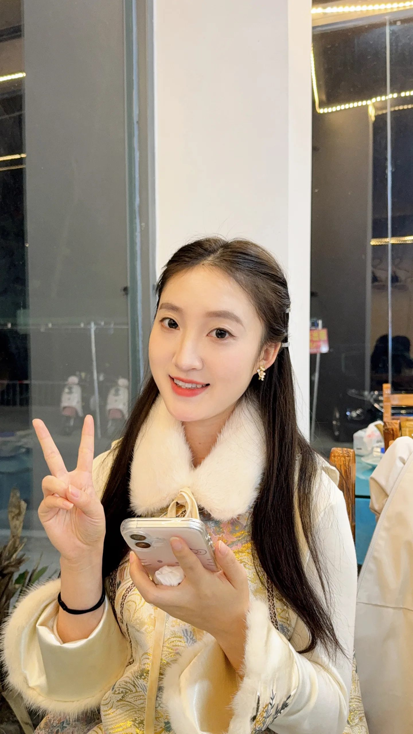 图片展示了一位年轻女性，她面带微笑，右手比出“V”字手势，左手持手机。她穿着白色毛领外套，内搭黄色花纹上衣，耳戴耳环。背景为室内环境，左侧有玻璃隔断，右侧有