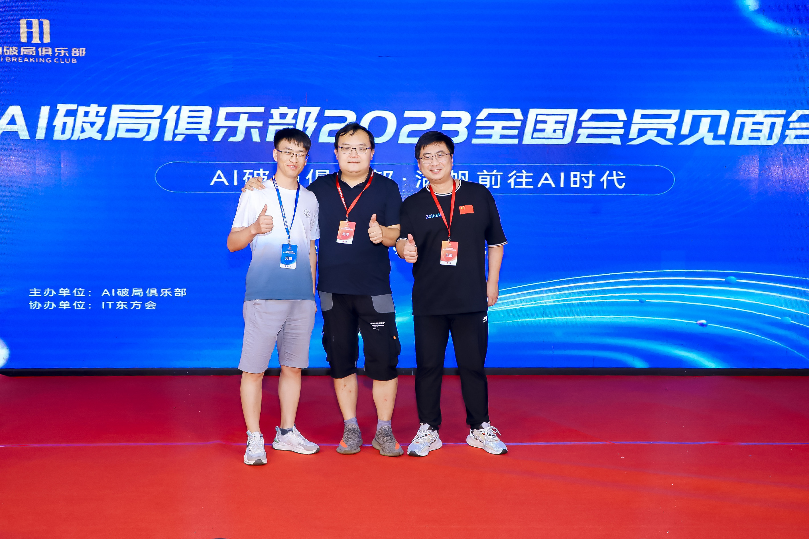 图片展示了AI破局俱乐部2023全国会员见面会现场，三位男士站在红色地毯上，背景为蓝色带有水波纹的屏幕，屏幕上方有“AI破局俱乐部2023全国会员见面会”及“AI破局俱乐部 汇聚前往AI时代”的字样。左侧男士穿浅色短袖和短裤，中间男士穿深色短袖和短裤，右侧男士穿黑色长袖和长裤。他们面带微笑，竖起大拇指，显得非常开心。此图与上文提到的作者在北京参加AI破局线下大会相呼应，体现了作者在大会中的美好体验。