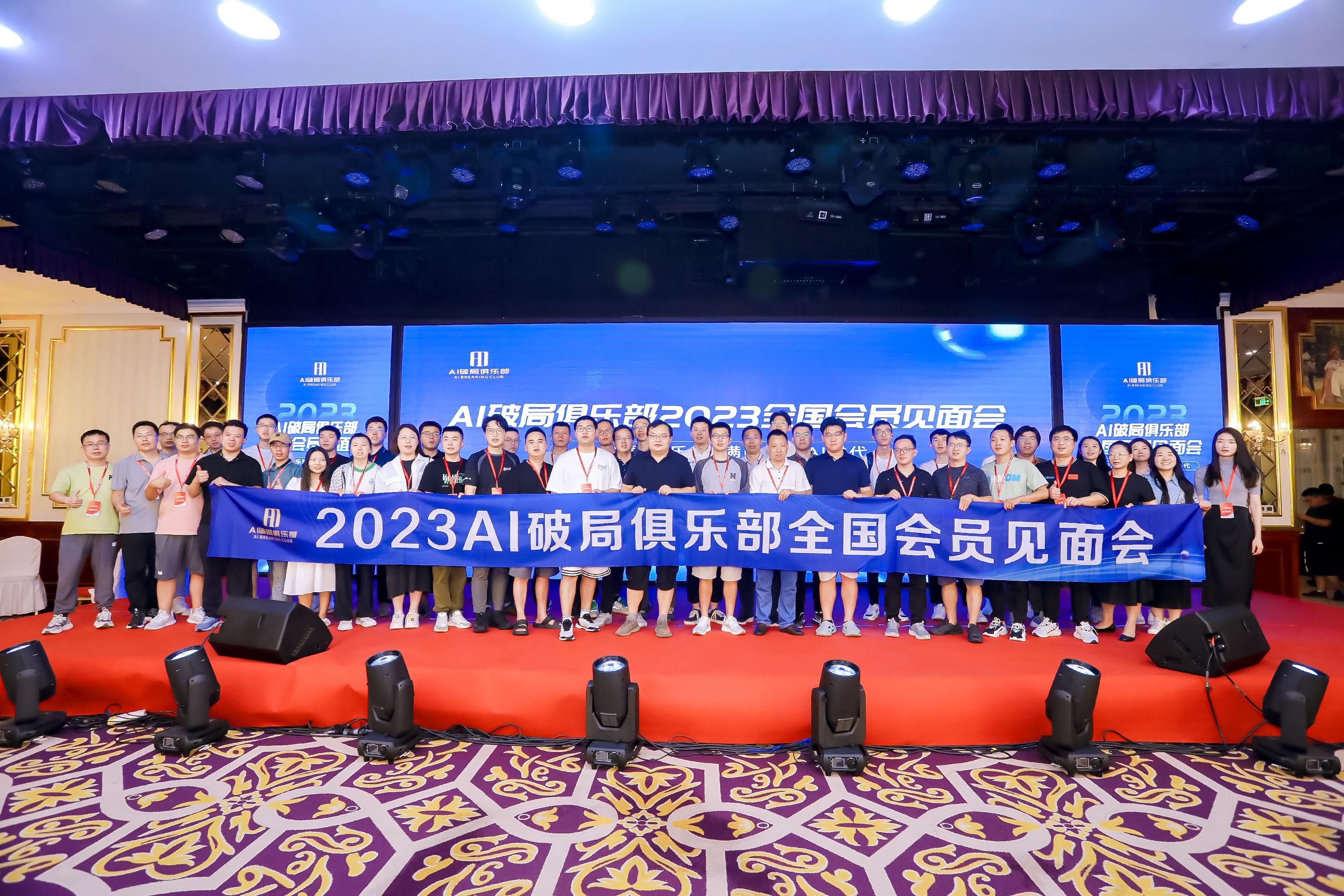 图片为2023AI破局俱乐部全国会员见面会现场合影。背景有紫色幕布和蓝色屏幕，屏幕上显示“2023 AI破局俱乐部全国会员见面会”字样。台前有红色地毯，多人站成两排，手持写有“2023AI破局俱乐部全国会员见面会”字样的蓝色横幅。现场灯光充足，氛围热烈。此图与上文提到的北京线下大会和潘神、玉润等伙伴面基相呼应，展示了线下交流的场景。