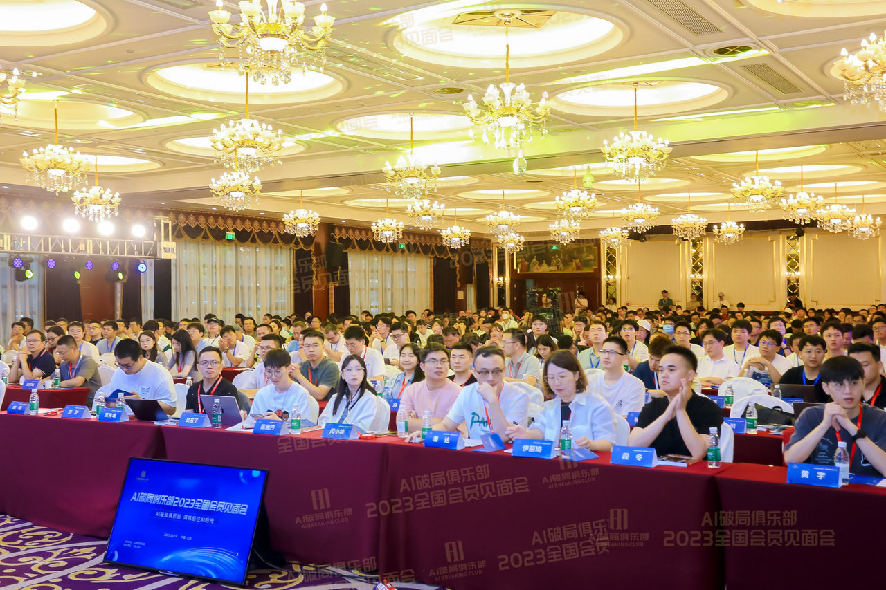 图片展示的是2023年AI破局俱乐部全国会员见面会现场。会场内装饰华丽，悬挂着多盏吊灯，背景有“AI破局俱乐部 2023全国会员见面会”字样。台前摆放着红色桌布，上面印有相同标识，台上有水瓶、麦克风等物品。台前坐着多位身着白色上衣的工作人员，部分人佩戴着工作证。台前还设有电脑显示屏，显示着相关内容。此图与上文提到的破局俱乐部全国见面大会相呼应，直观呈现了活动场景。