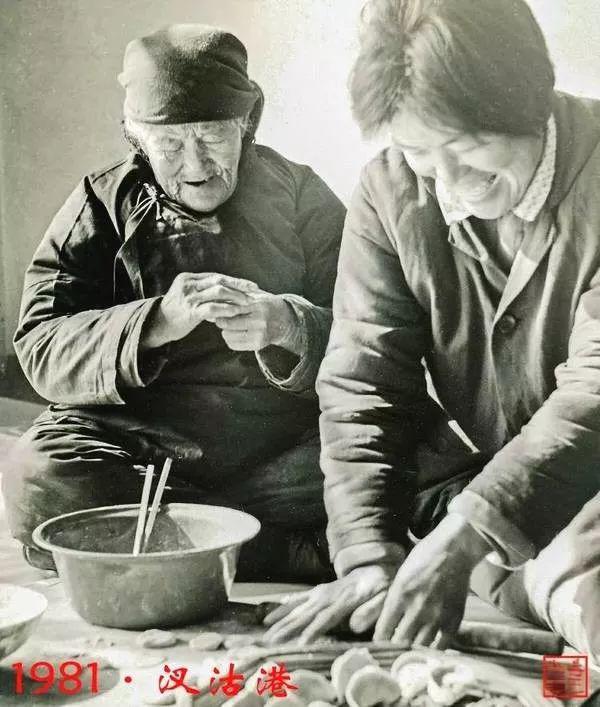 图片展示了一张1981年汉沽港的老照片，画面中有两位女性，一位坐在地上，另一位站着，她们正在制作食物。坐着的女性穿着深色上衣，戴着帽子，手里拿着筷子，面前有一个装有食物的碗；站着的女性穿着浅色上衣，正在擀面。照片左下角有“1981·汉沽港”的字样，右下角有一个红色印章。该图片用于展示修复前的效果，与文档中介绍的AI老照片修复操作上下文相关，可对比修复后的效果。