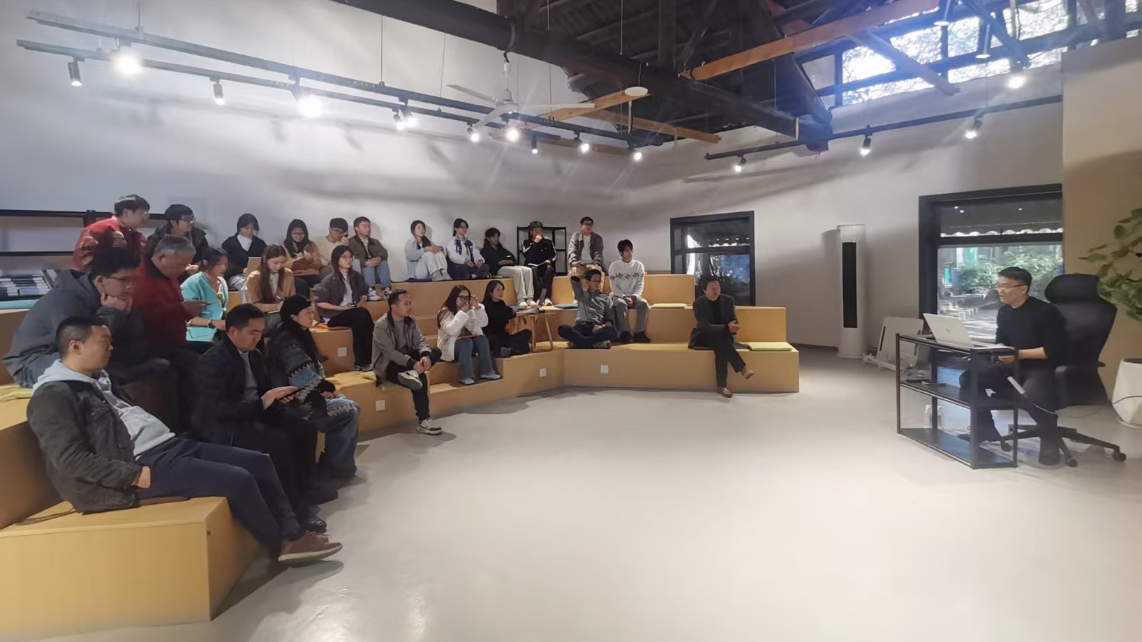 图片展示了一场N8N专题培训的场景。培训地点在一个室内空间，天花板上有排列整齐的射灯。左侧有几位坐在黄色沙发上的学员，他们或专注听讲，或低头记录。右侧有一位坐在黑色办公椅上的讲师，他手持文件，正在讲解。背景中还有几扇窗户，窗外景色隐约可见。此图与文档中“昨天搞了一场N8N专题培训”的内容相契合，直观呈现了培训现场情况。
