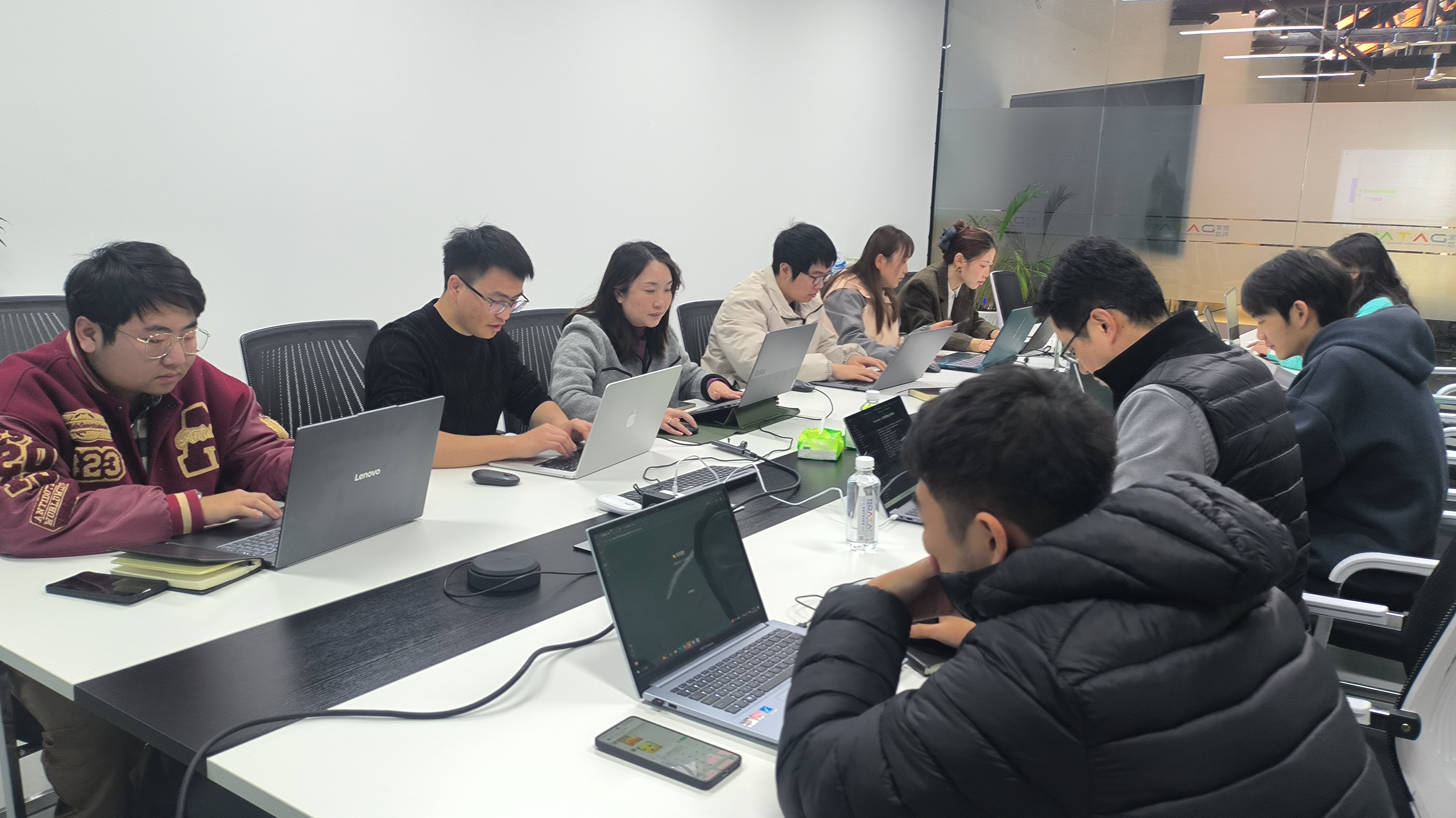 图片展示的是昨天培训现场的场景。多位学员围坐在长桌旁，专注地使用笔记本电脑。桌面上摆放着水瓶、手机等物品。背景中可以看到墙上的装饰和一些植物。这张图片与文档中“昨天培训现场，我发现了小白学N8N的三大死穴”的内容相呼应，直观呈现了培训现场学员学习N8N工作流的场景，为描述小白学N8N时可能遇到的困难提供了直观的背景。