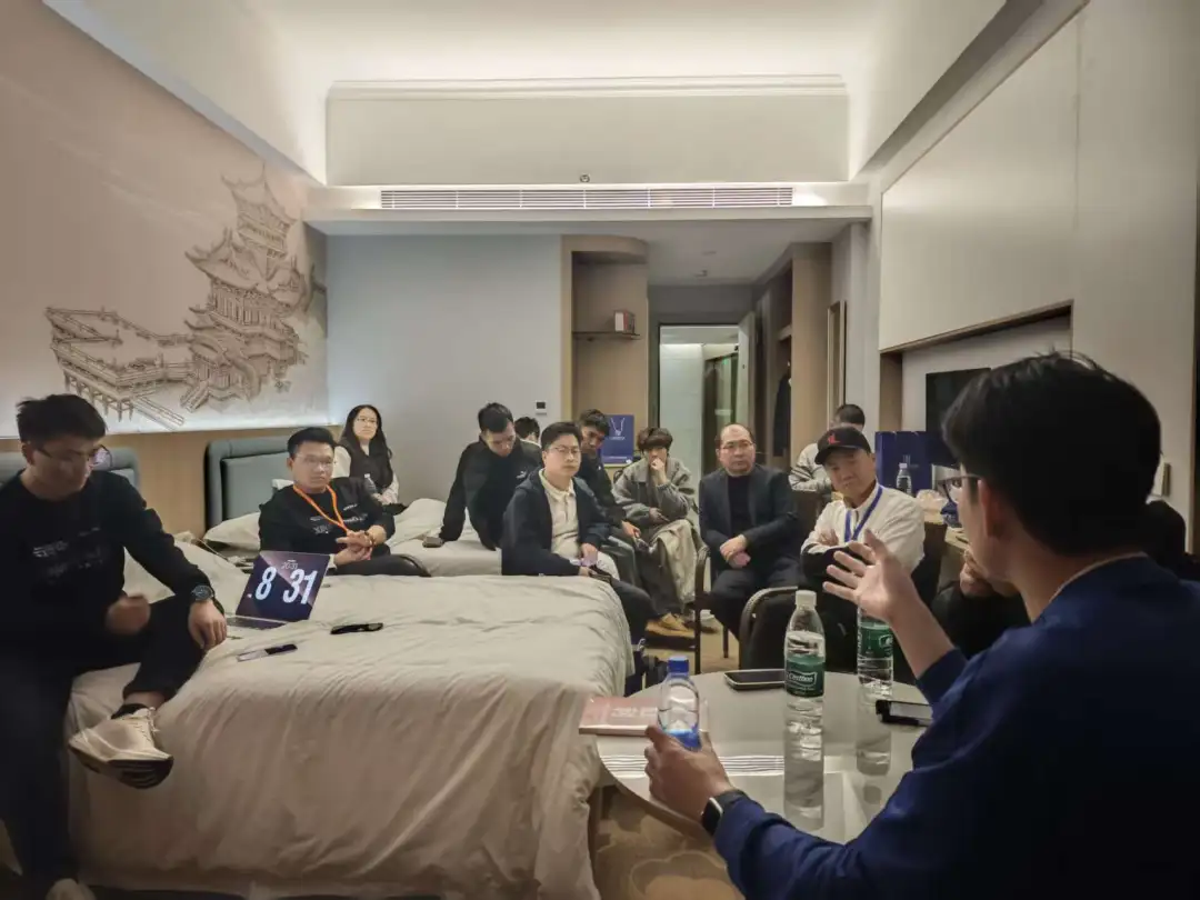 图片展示的是一个室内场景，多人围坐在一张铺有白色床单的床边，似乎在进行交流。床上有笔记本电脑、水瓶等物品。房间内有空调出风口、门等设施，墙上挂着一幅建筑图案。其中一位男士手持手机，另一位男士在用笔记本电脑演示。此场景与文档中“晚上夜话组局更有意思”相呼应，可能是文冠分享做IP经验、演示必火GEO营销系统全流程的场景。