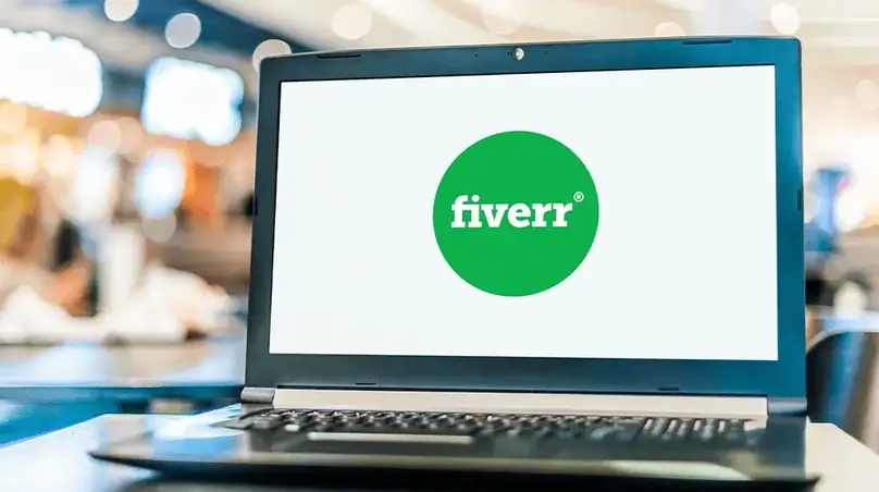 图片展示了一台笔记本电脑，屏幕上显示着Fiverr平台的标志，标志由一个绿色圆圈和白色“fiverr”字样组成。背景模糊，隐约可见室内环境。该图片与文档中介绍Fiverr平台的内容相关，直观呈现了Fiverr的标志，辅助说明Fiverr作为国际化的众包平台，为全球用户提供多种多样的服务这一背景信息。
