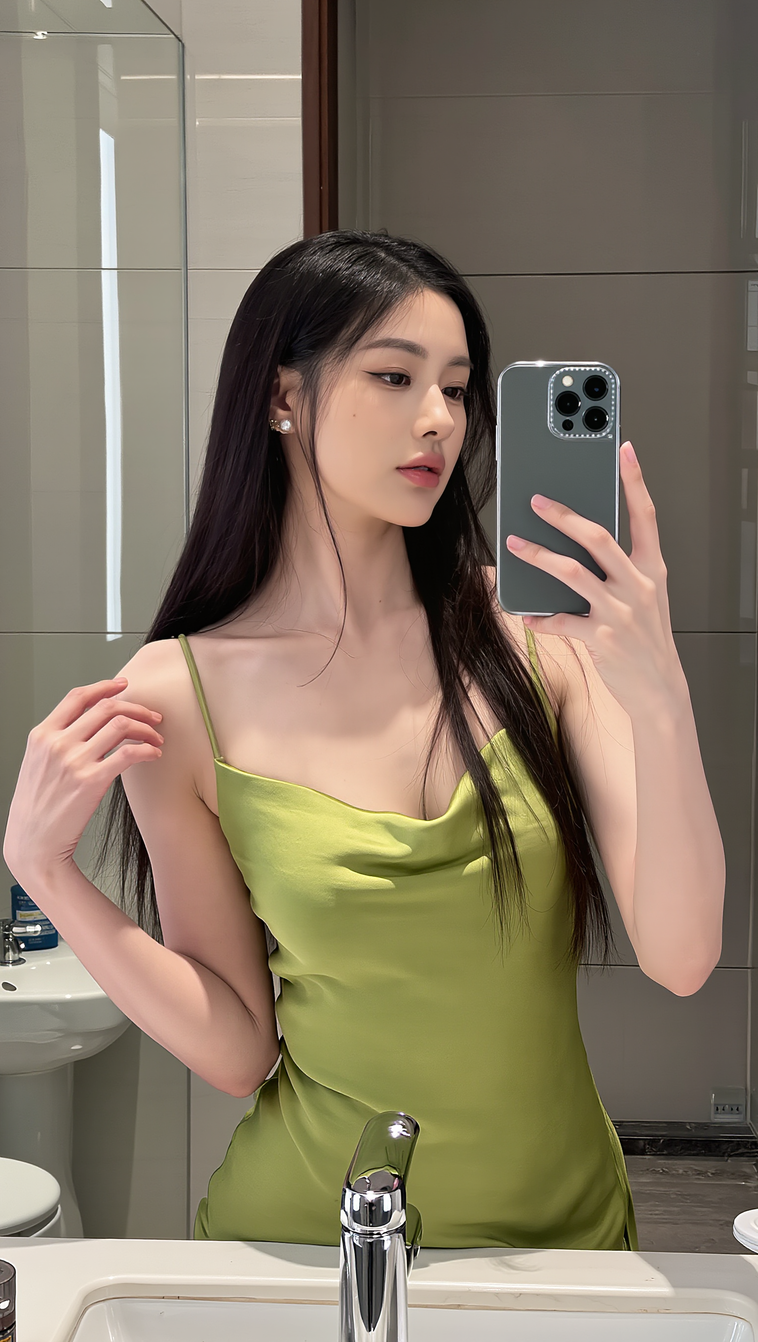图片展示了一位女性在浴室镜子前自拍的场景。她身着绿色吊带裙，长发披肩，左手持手机，右手自然下垂。背景中可见洗手台、水龙头、镜子及部分浴室设施。此图对应文档中“Step 1：上传你的照片”步骤，用以说明上传照片时脸部清晰、光线自然、表情自然等要求，直观呈现了上传照片的示例。