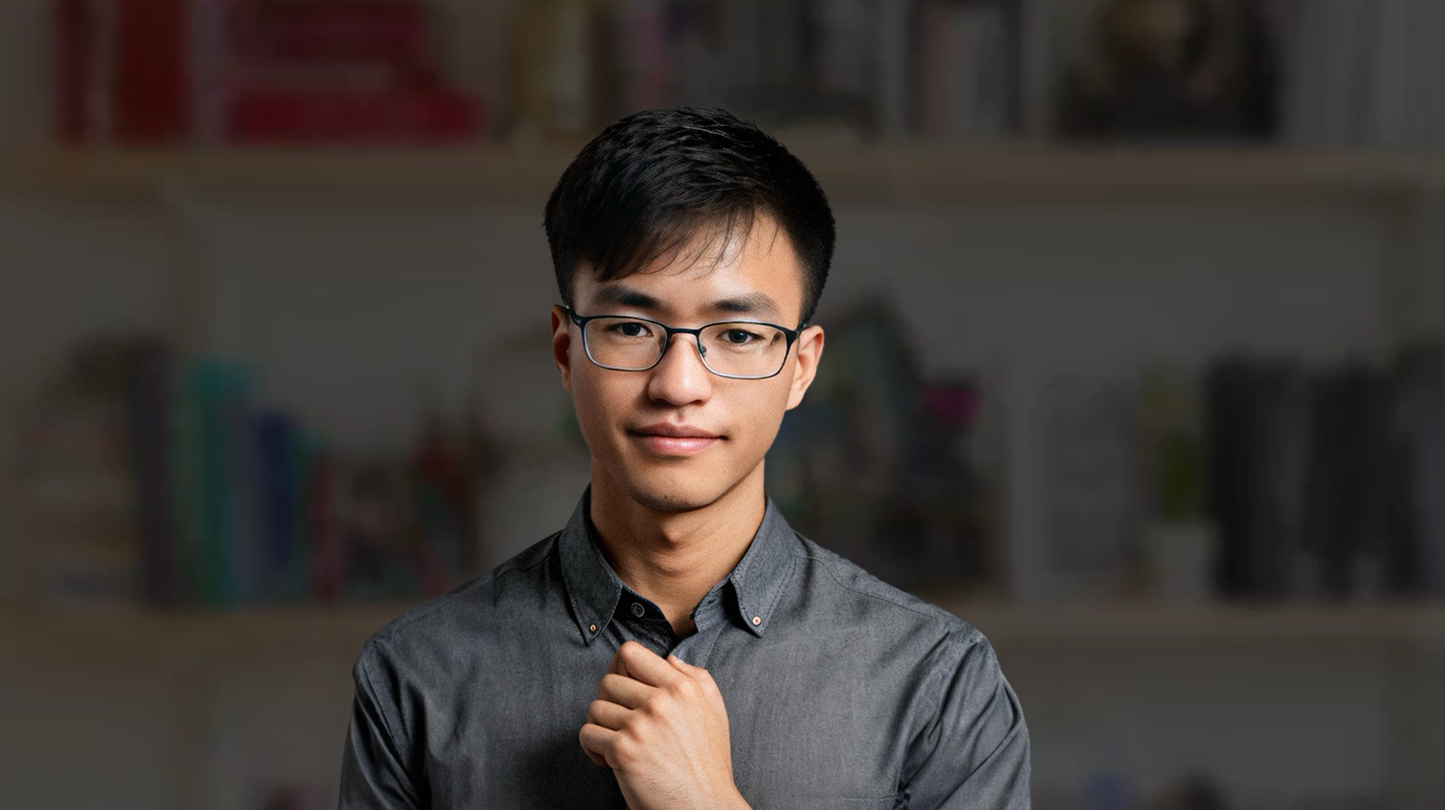图片展示的是一位戴着眼镜的男性形象，身穿灰色衬衫，单手放在胸前，神态自然。背景是一个书架，上面摆放着一些书籍和物品，画面整体较为清晰。这张图片是“元峰和安娜的形象图也都准备好了”中提到的元峰的形象图，是AI数字人访谈类视频幕后制作中准备的形象素材之一。