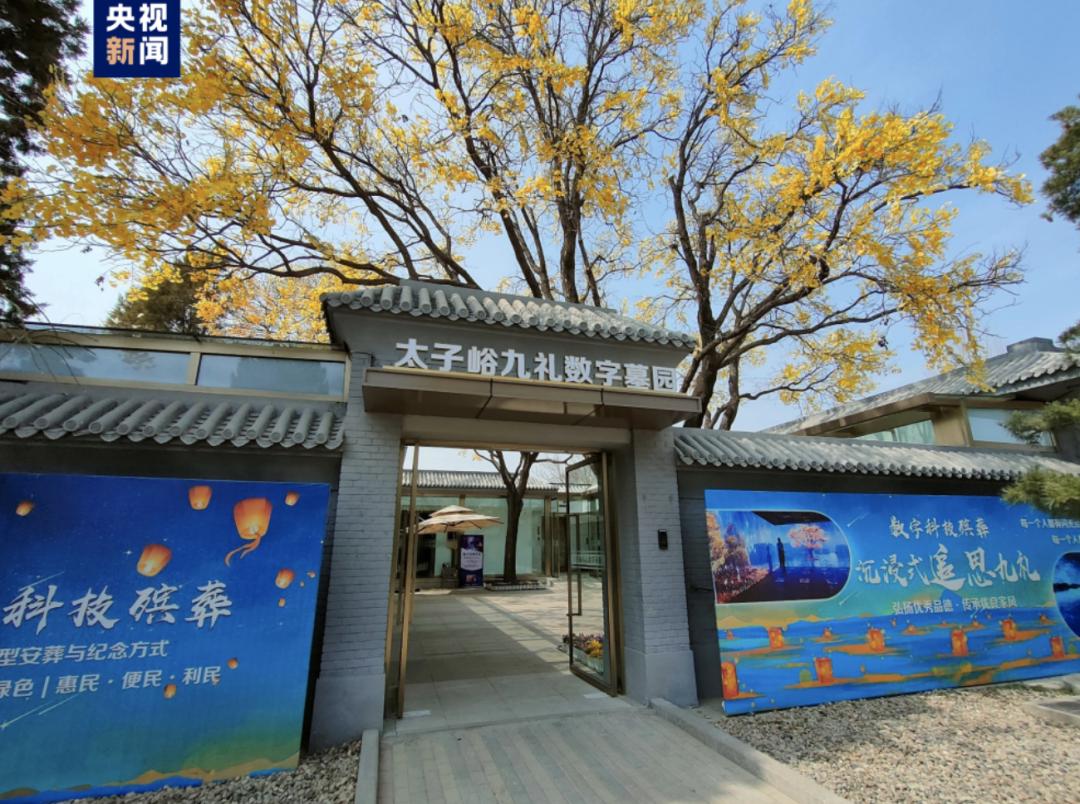 图片展示的是北京太子峪九礼数字墓园的入口。入口上方有“太子峪九礼数字墓园”的字样，两侧墙上分别有宣传海报，左侧写着“科技殡葬”等内容，右侧有“数字科技殡葬”等字样。墓园入口处有树木，树叶呈现出金黄色。该图片与上下文的关系是，作为北京数字墓园的实景呈现，直观展现了文中所提及的中国殡葬行业数字化革命中数字墓园的场景，为介绍北京数字墓位等相关内容提供了视觉参照。